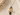 A gold-tone pendant necklace featuring a rectangular green stone, a white spiral shell charm, a faceted dark brown teardrop, and a clear flower charm with a purple center, displayed on a textured beige surface with dried flowers.