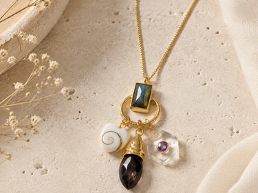 A gold-tone pendant necklace featuring a rectangular green stone, a white spiral shell charm, a faceted dark brown teardrop, and a clear flower charm with a purple center, displayed on a textured beige surface with dried flowers.