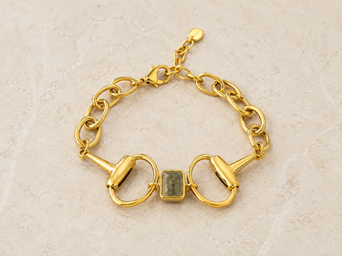 A gold horsebit chain bracelet with a clear square-cut gemstone in the center on beige table