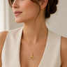 A woman wearing a delicate gold necklace with a small open oval pendant and a matching small gold stud earring, paired with a cream-colored sleeveless top.