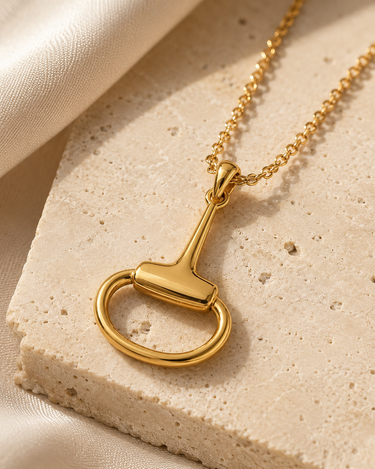 A polished gold horsebit pendant necklace on a delicate gold chain, styled on a textured beige stone slab with draped cream fabric.