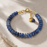 A beaded bracelet made of polished blue round beads with a gold-tone clasp and two charms, one heart-shaped and one teardrop-shaped dark blue stone, displayed on a textured beige stone surface.