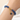 A close-up of a blue beaded bracelet with a gold clasp and dangling charm, worn on a person's wrist against a soft white background.