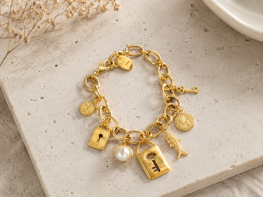 A gold charm bracelet with a link chain and multiple charms, including a keyhole, key, lock, fish, and a white pearl, displayed on a light beige stone surface.