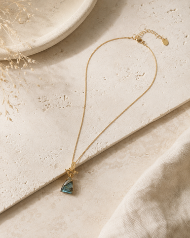 A delicate gold chain necklace with a small triangular blue-green stone pendant, styled on a textured beige stone slab with a soft cream fabric and dried grass in the background.