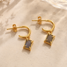 A pair of gold hoop earrings with rectangular gray stone pendants, displayed on a textured beige surface with dried flowers.
