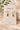 A pair of gold hoop earrings with rectangular green gemstones, displayed on a white brand card labeled CHAN LUU, set against a warm beige stone background with soft leafy props.