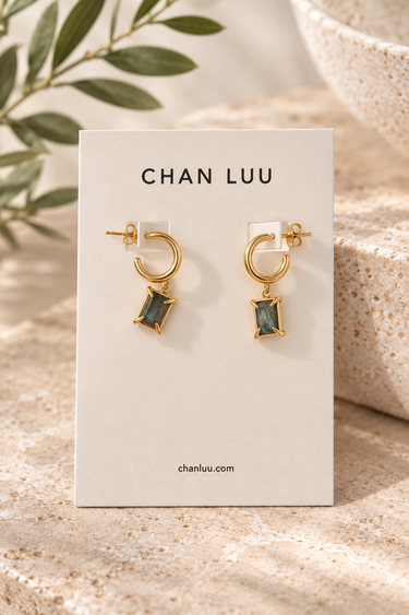 A pair of gold hoop earrings with rectangular green gemstones, displayed on a white brand card labeled CHAN LUU, set against a warm beige stone background with soft leafy props.