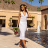 Woman in a white dress standing by a pool with palm trees and mountains in the background