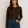 A woman wearing a dark brown long-sleeve cropped cardigan with a button-down front and blue jeans, standing against a plain light gray background.