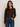 A woman wearing a dark brown long-sleeve cropped cardigan with a button-down front and blue jeans, standing against a plain light gray background.