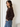 A woman wearing a dark brown knit cardigan sweater and blue jeans, shown in profile against a neutral off-white wall.