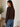 A woman wearing a dark brown long-sleeve knit sweater and blue jeans, shown from the back in a softly lit indoor setting with a wooden table and potted plant in the background.