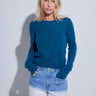 A woman modeling a teal blue long-sleeve knit sweater with light-wash blue jeans against a light gray studio background.