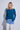 A woman modeling a teal blue long-sleeve knit sweater with light-wash blue jeans against a light gray studio background.