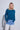 A blonde woman modeling a teal blue long-sleeve knit sweater with light-wash jeans against a light gray studio background.