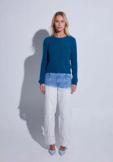 A woman stands against a light gray studio background wearing a teal long-sleeve knit sweater, a light blue denim waistband detail, white wide-leg cropped jeans, and light blue pointed heels.