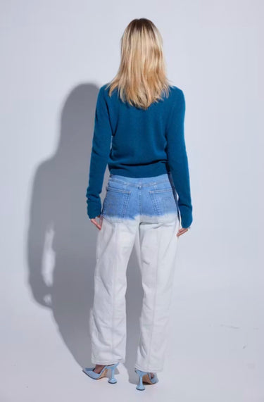 A woman is shown from behind wearing a teal blue long-sleeve knit sweater, light-wash distressed denim jeans, and light blue heels, standing against a light gray studio background with a prominent shadow.