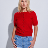 A blonde woman modeling a short-sleeve red knit cardigan with front buttons, paired with light blue jeans and white pants, standing against a light gray studio background.