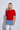 A blonde woman modeling a short-sleeve red knit cardigan with front buttons, paired with light blue jeans and white pants, standing against a light gray studio background.