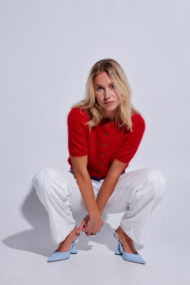 A blonde woman crouching in a studio setting, wearing a bright red short-sleeve button-front top, white wide-leg pants, and light blue pointed-toe heels.