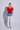 A woman stands with her back to the camera wearing a bright red short-sleeve top, light blue and white washed denim jeans, and blue sneakers against a light gray studio background.