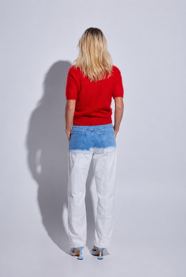 A woman stands with her back to the camera wearing a bright red short-sleeve top, light blue and white washed denim jeans, and blue sneakers against a light gray studio background.