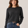 A woman wearing a dark charcoal gray knit crewneck sweater and blue jeans, standing against a plain light beige studio background.