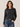 A woman wearing a dark charcoal gray knit crewneck sweater and blue jeans, standing against a plain light beige studio background.