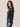 A woman wearing a dark charcoal gray knit crewneck sweater and blue jeans, standing against a plain light beige studio background.
