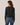A woman shown from the back wearing a dark charcoal gray knit sweater and blue jeans, standing against a plain light beige studio background.