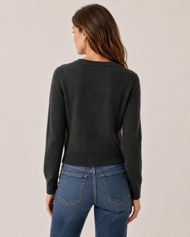 A woman shown from the back wearing a dark charcoal gray knit sweater and blue jeans, standing against a plain light beige studio background.