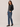 A woman modeling a dark charcoal gray long-sleeve knit sweater and blue straight-leg jeans with black loafers, standing against a solid light beige studio background.