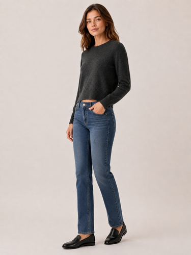 A woman modeling a dark charcoal gray long-sleeve knit sweater and blue straight-leg jeans with black loafers, standing against a solid light beige studio background.