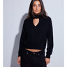A woman with long dark brown hair wearing a black knit mock neck sweater with a heart-shaped keyhole cutout, photographed against a light gray studio background.