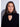 A woman with long dark brown hair wearing a black knit mock neck sweater with a heart-shaped keyhole cutout, photographed against a light gray studio background.