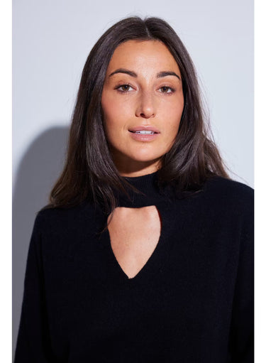 A woman with long dark brown hair wearing a black knit mock neck sweater with a heart-shaped keyhole cutout, photographed against a light gray studio background.