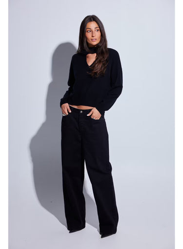 A woman with long dark brown hair wearing a black knit mock neck sweater with a heart-shaped keyhole cutout, and black jeans photographed against a light gray studio background.