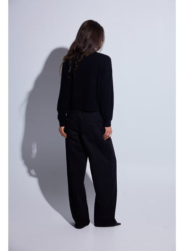 A woman with long dark brown hair wearing a black knit mock neck sweater with black jeans photographed against a light gray studio background.