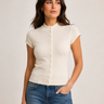 A woman wearing a cream-colored short-sleeve knit top with a button-down front and a high mock neck, paired with blue jeans, against a plain light gray background.