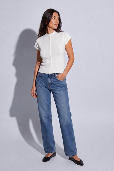 A woman modeling a white short-sleeve button-front top and blue straight-leg jeans against a light gray studio background.