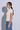 A woman in side profile wearing a fitted short-sleeve cream knit top and blue jeans, standing against a light gray studio background with a soft shadow.