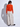 Woman facing backwards wearing a bright orange crewneck sweater layered over a white mock-neck top with relaxed light beige wide-leg pants.