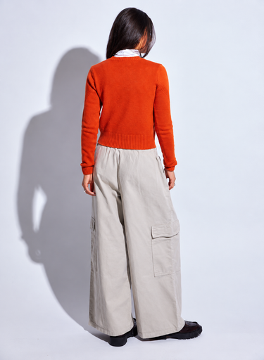 Woman facing backwards wearing a bright orange crewneck sweater layered over a white mock-neck top with relaxed light beige wide-leg pants.