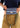 A person wearing a navy shirt and khaki pants holds a blue striped clutch bag with a colorful striped strap and a small gold-tone keychain. The bag has a tan leather brand patch that reads 'CONSUELA.