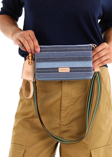 A person wearing a navy shirt and khaki pants holds a blue striped clutch bag with a colorful striped strap and a small gold-tone keychain. The bag has a tan leather brand patch that reads 'CONSUELA.