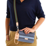 A person wearing a navy shirt and khaki pants holds a blue striped clutch bag with a colorful striped strap and a small gold-tone keychain. The bag has a tan leather brand patch that reads 'CONSUELA.