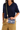 A person wearing a navy shirt and khaki pants holds a blue striped clutch bag with a colorful striped strap and a small gold-tone keychain. The bag has a tan leather brand patch that reads 'CONSUELA.