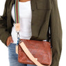 A woman wearing an olive green shirt and blue jeans carries a brown embossed leather crossbody bag with a beige strap and a colorful striped shoulder strap detail.
