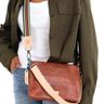 A woman wearing an olive green shirt and blue jeans carries a brown embossed leather crossbody bag with a beige strap and a colorful striped shoulder strap detail.